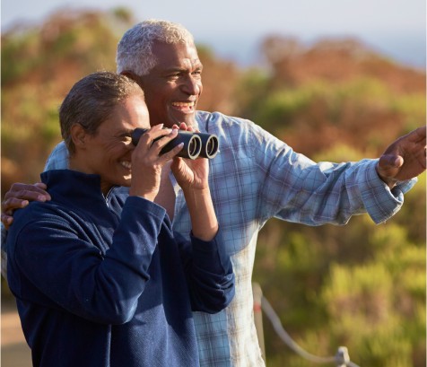 Two retired adults in a fall setting: a female looking through binoculars and a male gesturing towards something in the distance, with colorful autumn leaves in the background. 