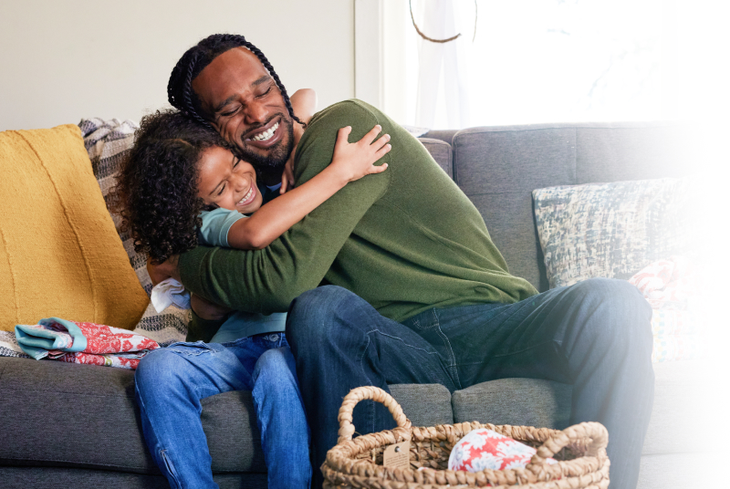 Father and young child sharing a joyful hug on a couch at home, symbolizing the personal impact of dental benefits.