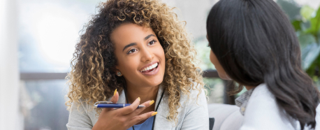 Two women engaged in a conversation, one holding a pen, illustrating broker-client collaboration.